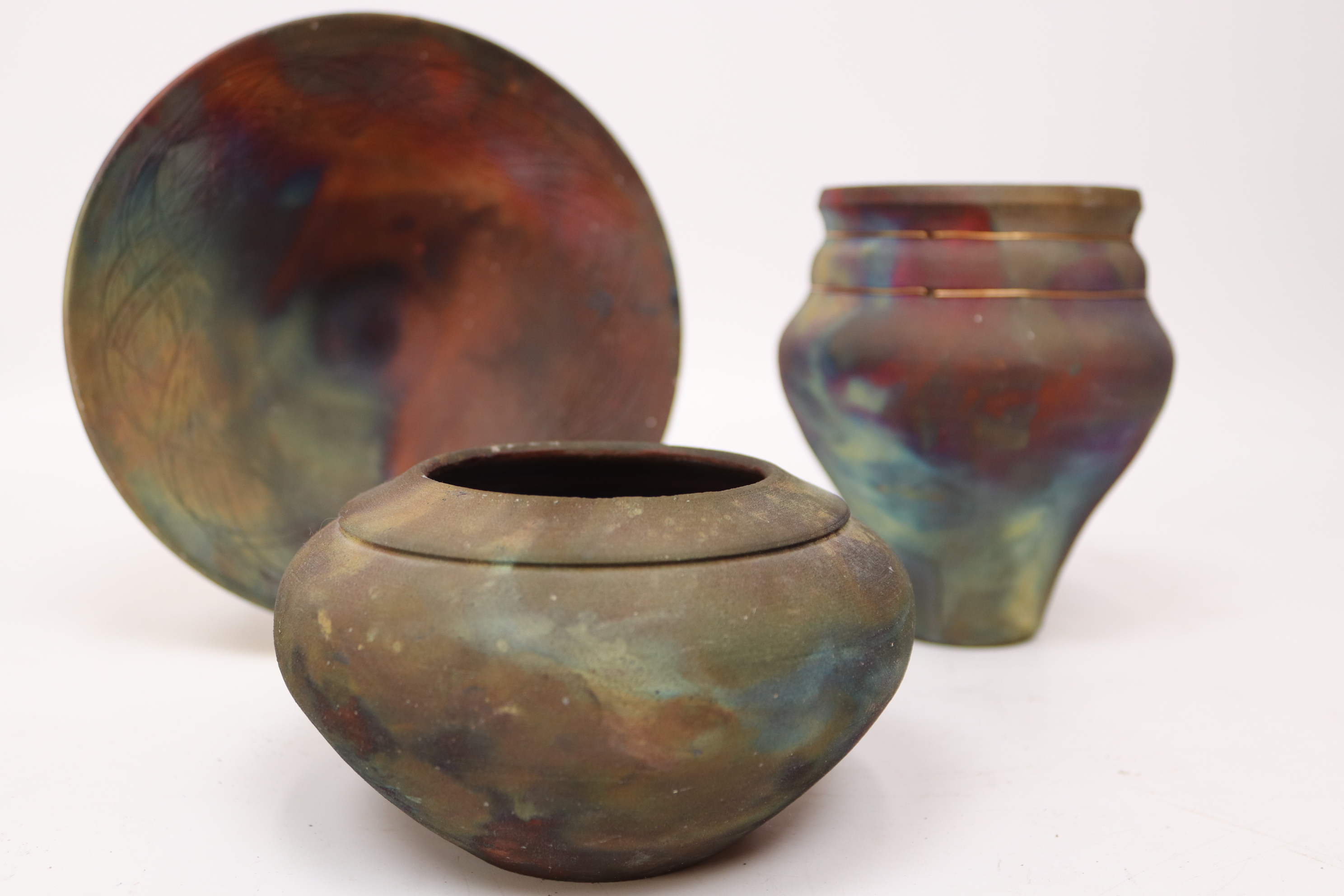 Three uniquely shaped pottery pieces with vibrant, swirling colors, including a shallow bowl and two jars, set against a plain background.